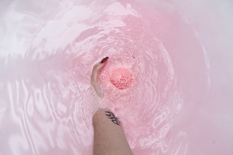 Model holding pink Awaken bath bomb in tub with pink water in background.