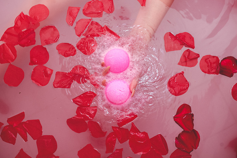 Model holds two pink Awaken CBD Kush Queen bath bombs in tub of water with floating rose petals.