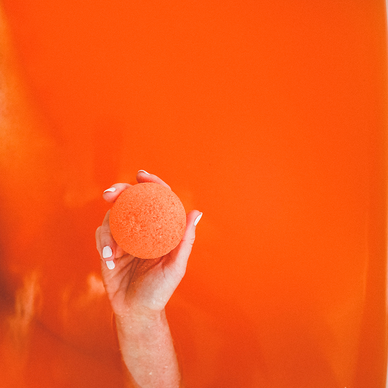 Model holds orange Citrus CBD bath bomb out of orange bath tub water.